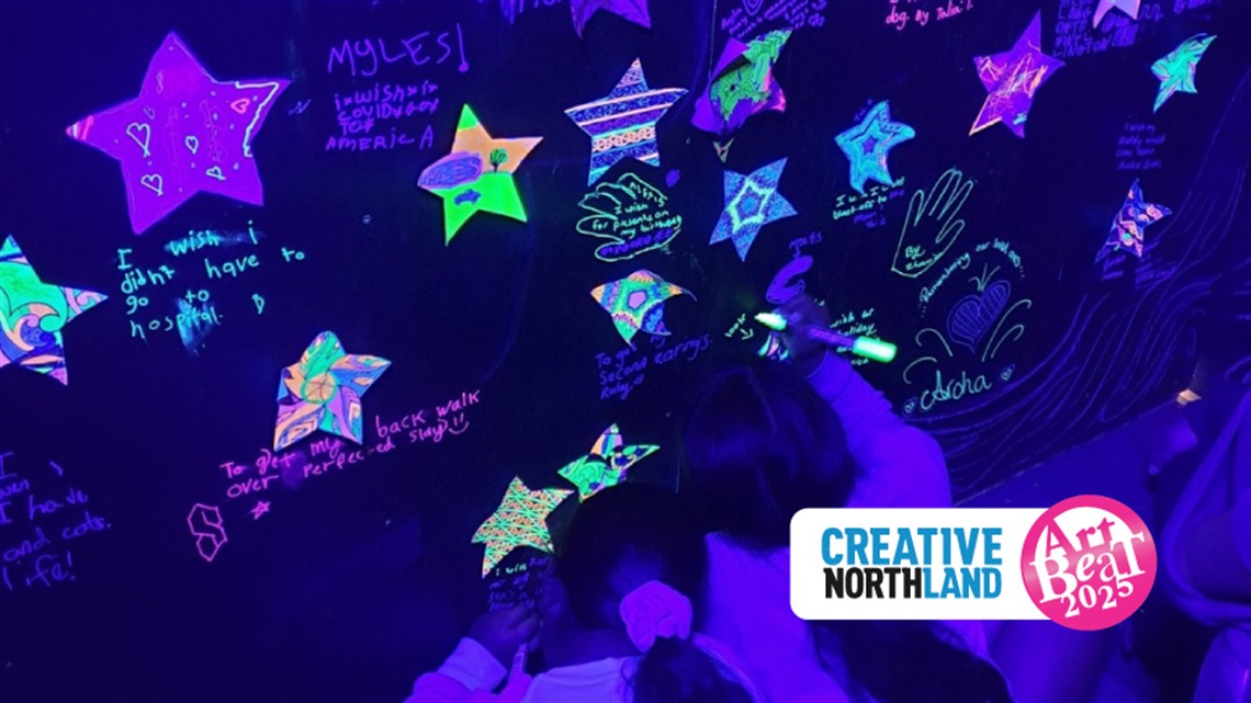Two children drawing on the wall of the glow room using irridescent pencils, with the Creative Northland ArtBeat 2025 logo. 