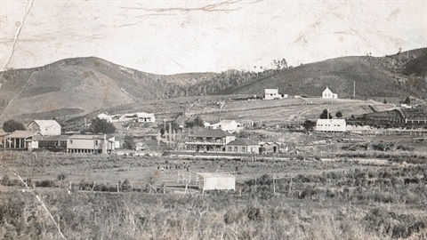 Photo Hukerenui in the 1926 showing a cluster of buildings, fields and grassy hills. 