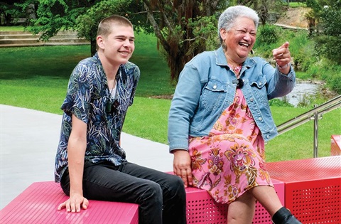 A young man and an older woman sit together on a bench in a park. 