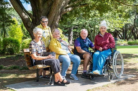 A few older people sitting on a bench in a park with a woman in a wheelchair. 