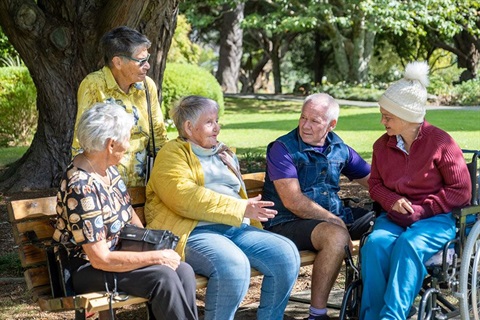 A group of older adults sitting on a park bench and in a wheelchair. 