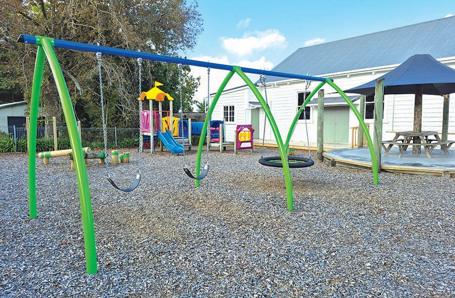 Children’s playground with swings, a slide and climbing equipment on a bark-covered play area beside a community building and shaded outdoor seating area. 