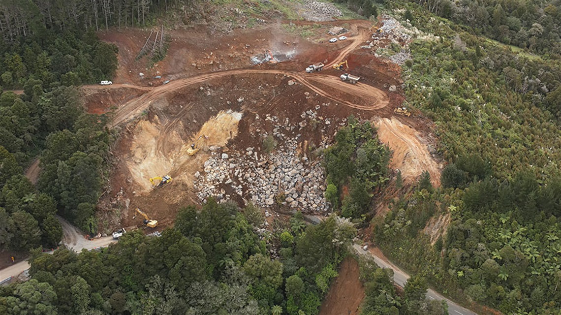 Aerial progress-photo of the slip at Helena Bay Hill from April 2026. The image shows heavy machinery clearing hundreds of large rocks and dark red soil. 