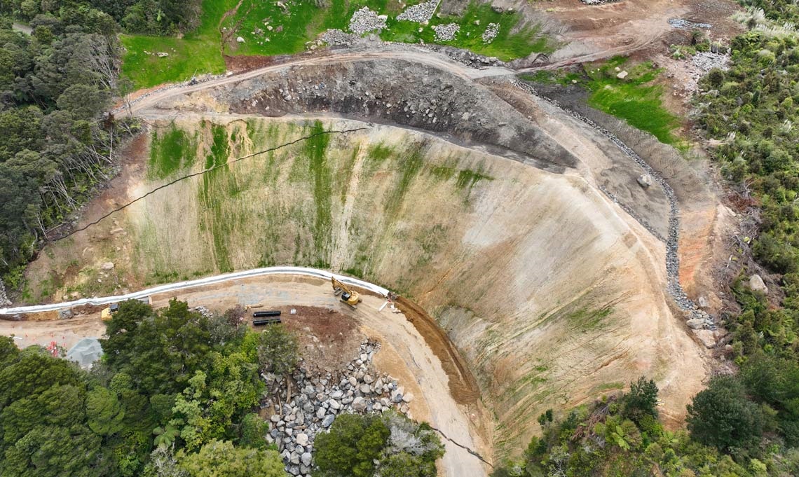 Aerial photo showing the enormous slip work-site on Russell Road at Helena Bay. Grass is starting to show on the treated slopes, and there is a gravel road where drainage work is taking place. 