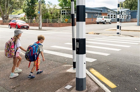 A young boy and girl wearing school bags on their backs approach a pedestrian crossing. 
