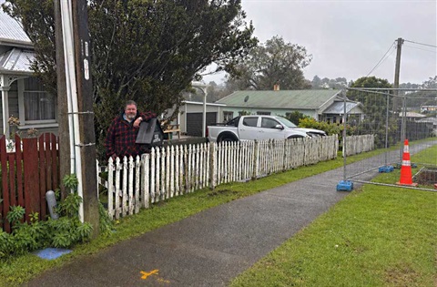 A man stands on his property behind a white picket fence with the pipe replacement works happening on his street. 