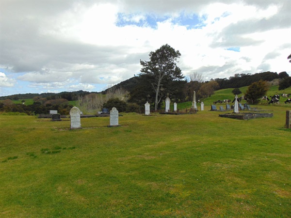 Mangapai Cemetery with weathered headstones on a grassy slope, surrounded by trees and open countryside under a cloudy sky. 