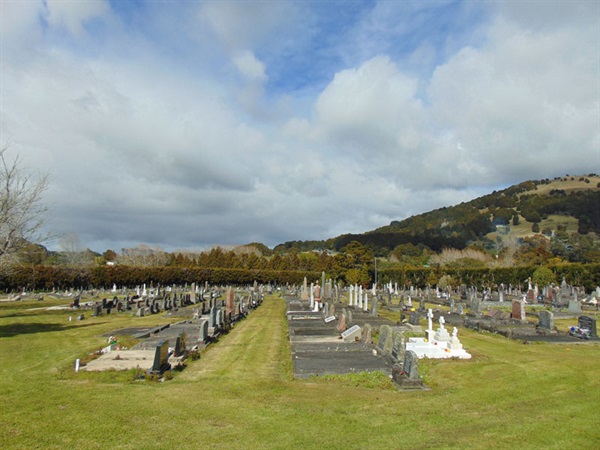 Gravestones at Kaurihohore Cemetery on a grassy lawn, with trees and a gentle hill rising in the background under a cloudy sky. 