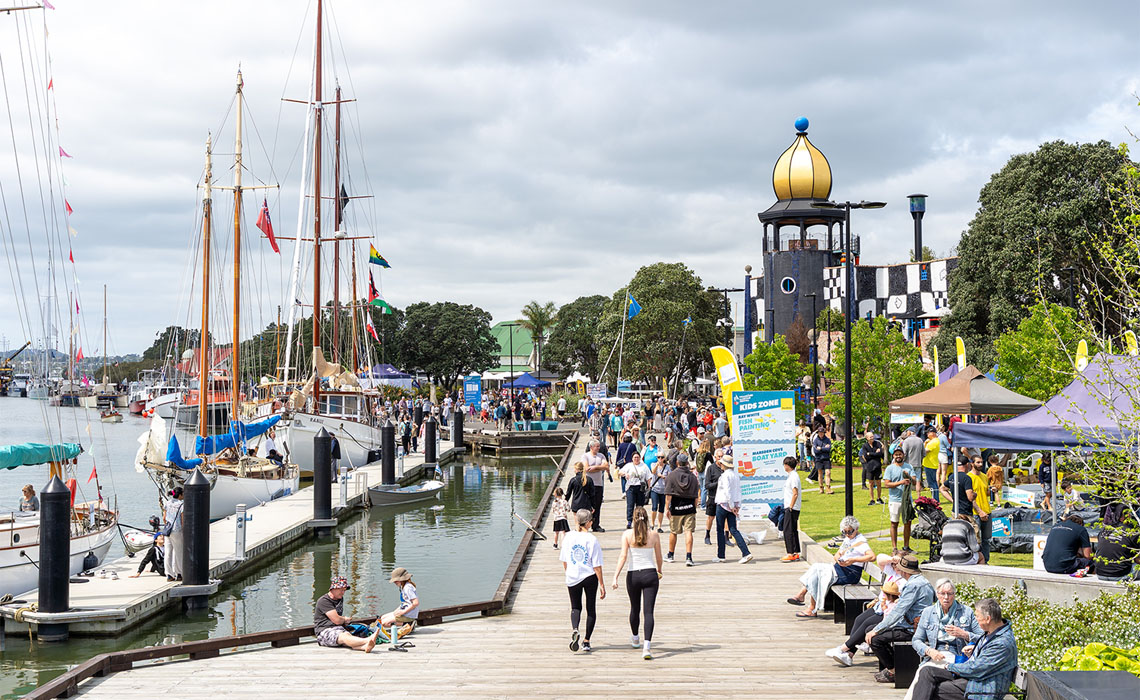 Crowds explore Whangārei’s Town Basin waterfront with boats, market stalls, and the distinctive gold‑topped Hundertwasser Art Centre in the background. 