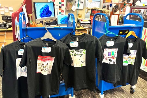 A row of custom-made black t‑shirts with hand‑drawn Te Tiriti o Waitangi designs hanging on a blue rolling rack in the library. 
