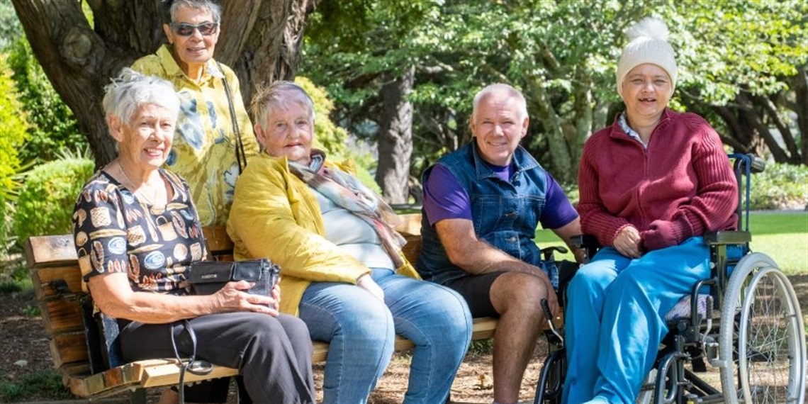 Five people sitting on a bench in an outdoor park setting, with one person in a wheelchair. The individuals' faces are obscured for privacy. The background features trees and greenery, suggesting a natural environment.