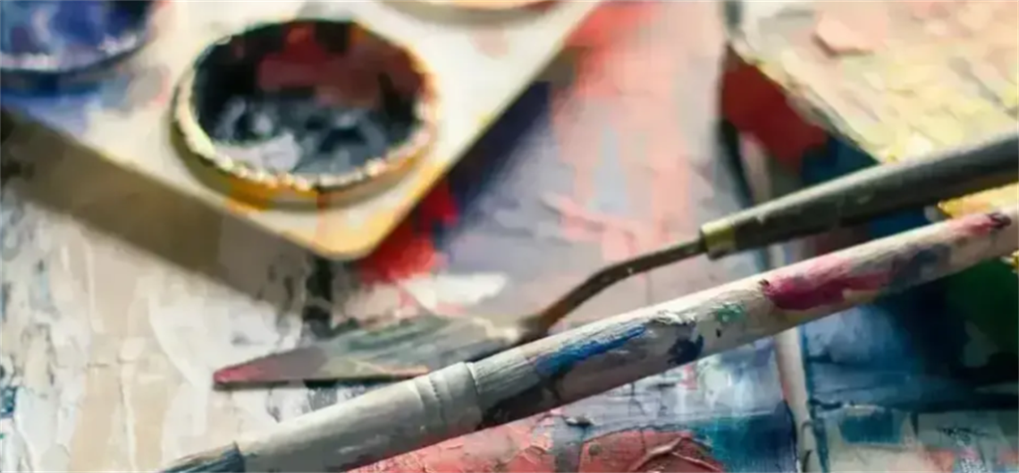Close-up of an artist's workspace with paintbrushes, a palette, and various painting tools on a surface covered in colorful paint splatters, reflecting an active and creative painting session.