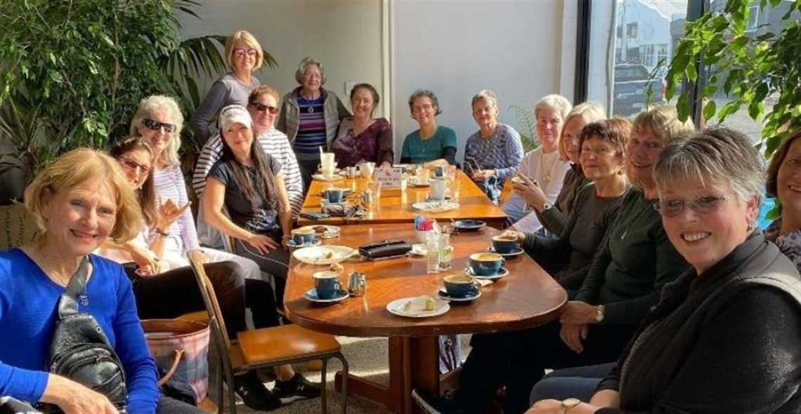 Group of people sitting around a long wooden table in a cafe or restaurant, with cups, plates, and dining items on the table. Background includes plants and large windows with natural light. Faces are pixelated for privacy. Group of people sitting around a long wooden table in a cafe or restaurant, with cups, plates, and dining items on the table. Background includes plants and large windows with natural light. Faces are pixelated for privacy.