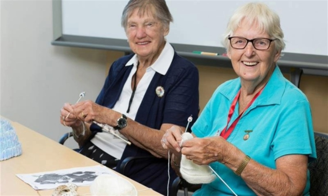 Two people sitting at a table indoors, knitting with yarn and needles, with a pattern sheet and balls of yarn in front of them.