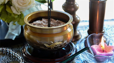 A memorial display featuring a brass incense burner filled with ash, lit incense sticks and a pink candle, with white flowers and decorative items arranged nearby.