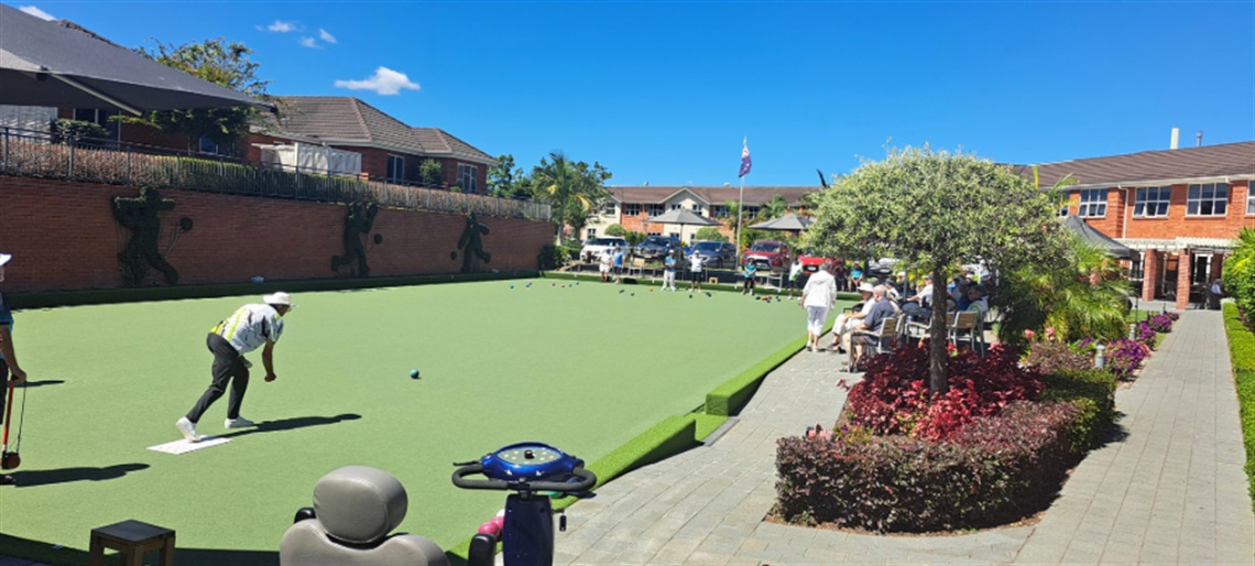 Outdoor lawn bowling green with players on the field and spectators seated under a shaded area, surrounded by buildings and landscaped greenery in a community setting.