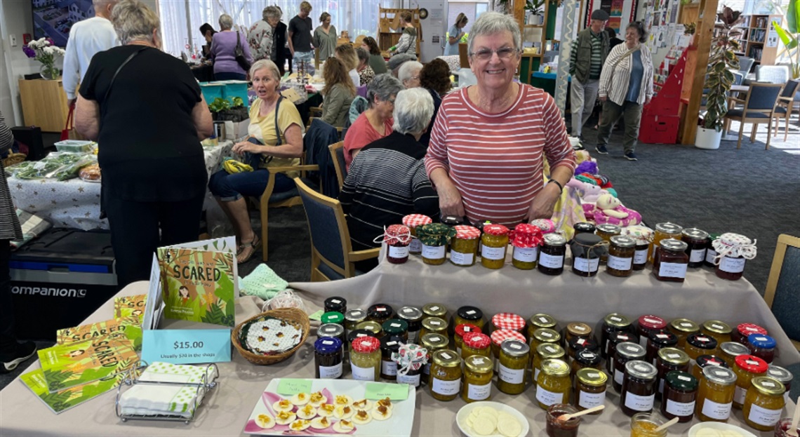 Indoor market scene at Summerset Mt Denby Market Day featuring stalls with homemade preserves, food samples, crafts, and a book titled 'SCARED' for sale, with attendees engaging in the lively event.