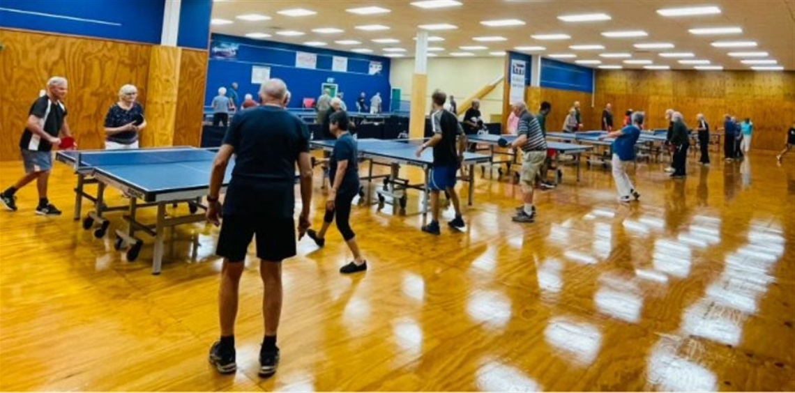 Indoor community table tennis event with multiple participants playing and socializing around several tables in a brightly lit hall with wooden floors and blue walls.