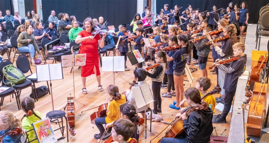 Children playing violins and cellos in a group music class or rehearsal, led by an instructor, with an audience watching in the background.