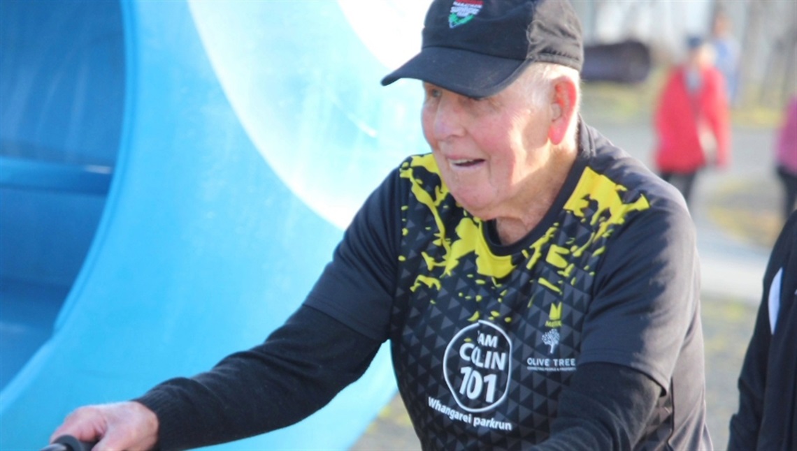 Elderly person using a walker, wearing a black cap and a black and yellow shirt with 'CLIM 707' text, in an outdoor setting with a large blue structure and other people in the background. Face is blurred for privacy.