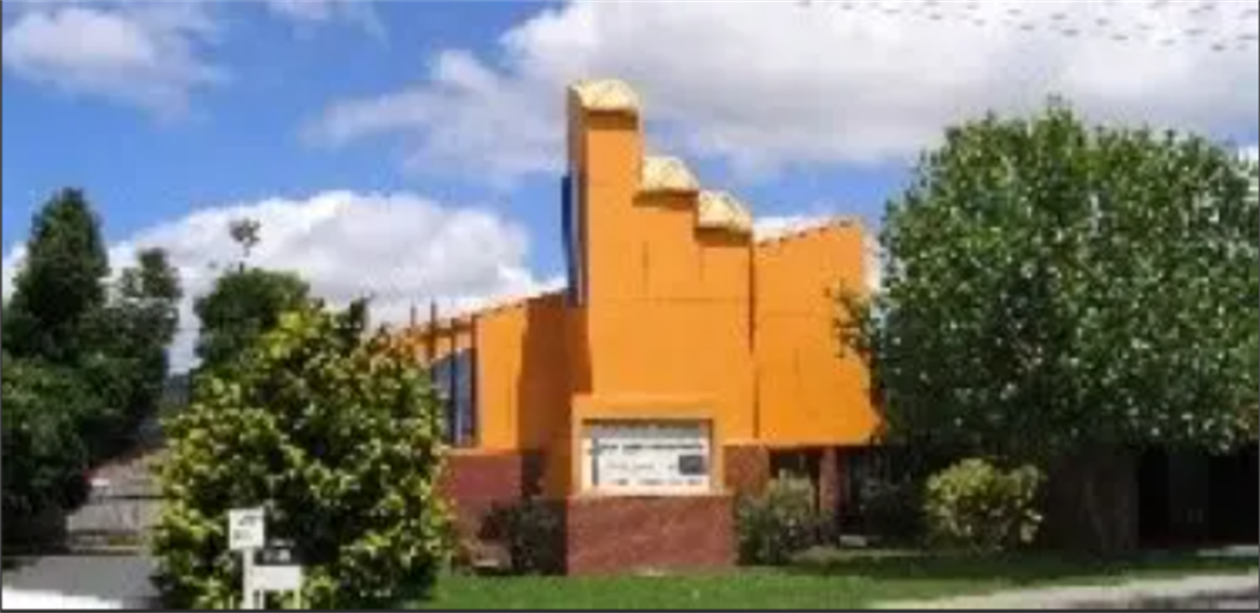 Bright orange building with three protruding roof structures, surrounded by trees and bushes. A sign is positioned in front of the building, though its text is unclear. The sky is partly cloudy with visible blue patches.