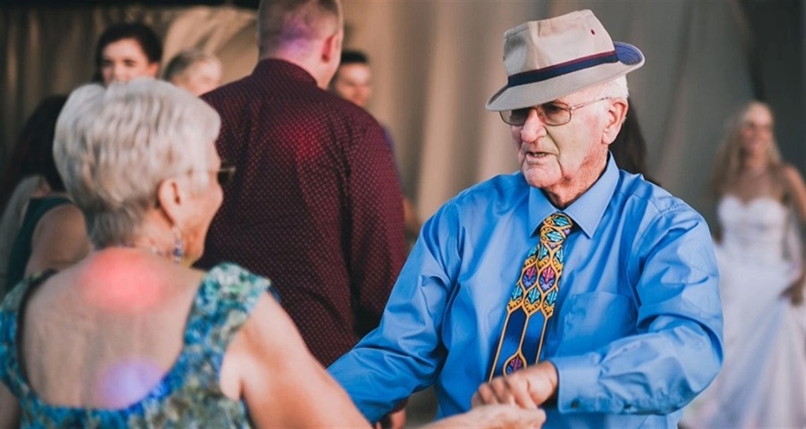 An older couple dancing joyfully at a social event. The man wears a blue shirt, colorful tie, and hat, while the woman wears glasses and a green-blue patterned sleeveless dress. Other attendees, including someone in a white dress, are visible in the background.