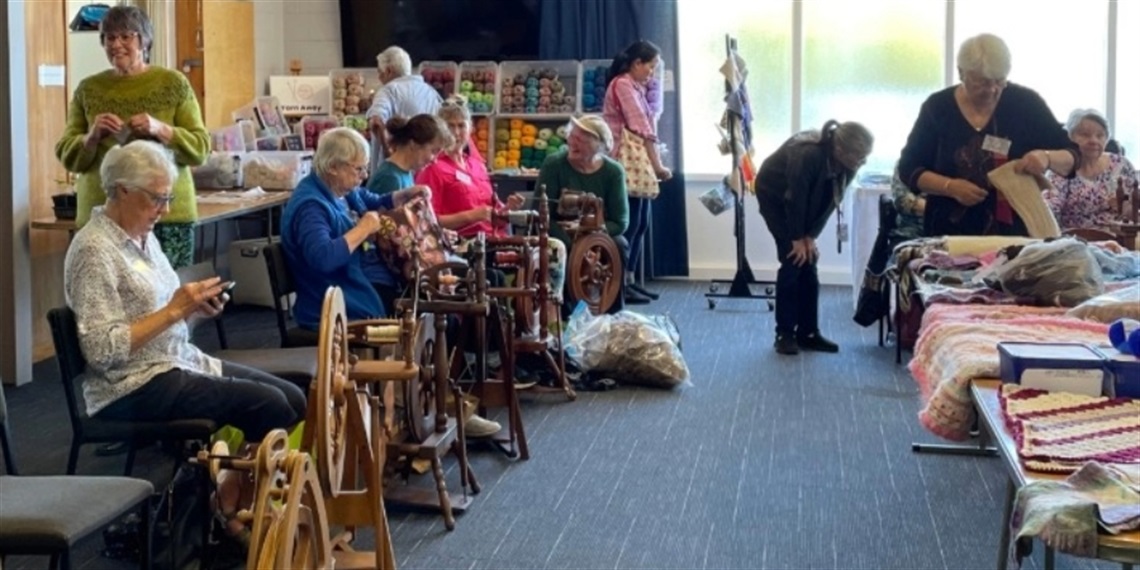 Group of people spinning yarn and displaying colorful knitted items in a communal textile craft space filled with yarn and supplies.