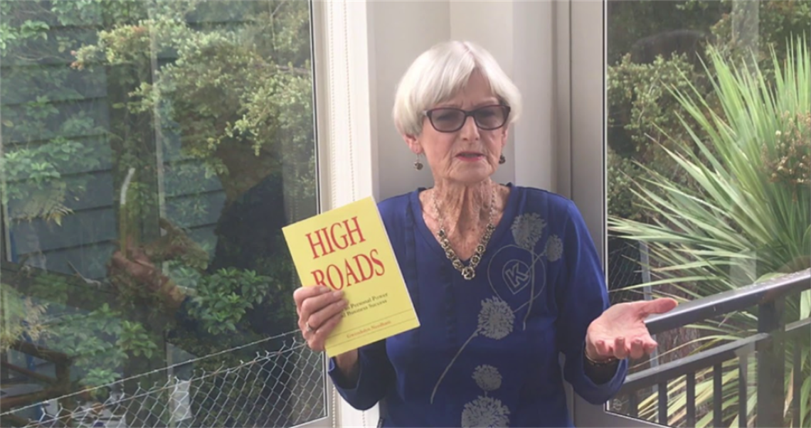 Person indoors holding a yellow book titled 'HIGH ROADS: A Journey Through Peace, Love and Happiness', with greenery visible through a large window in the background.