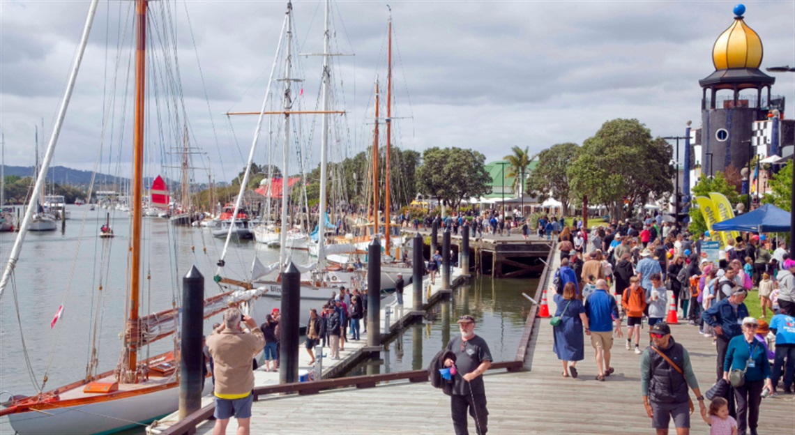 Crowds walking along a waterfront boardwalk during a maritime festival, with sailboats docked at the pier and a golden-domed building in the background.