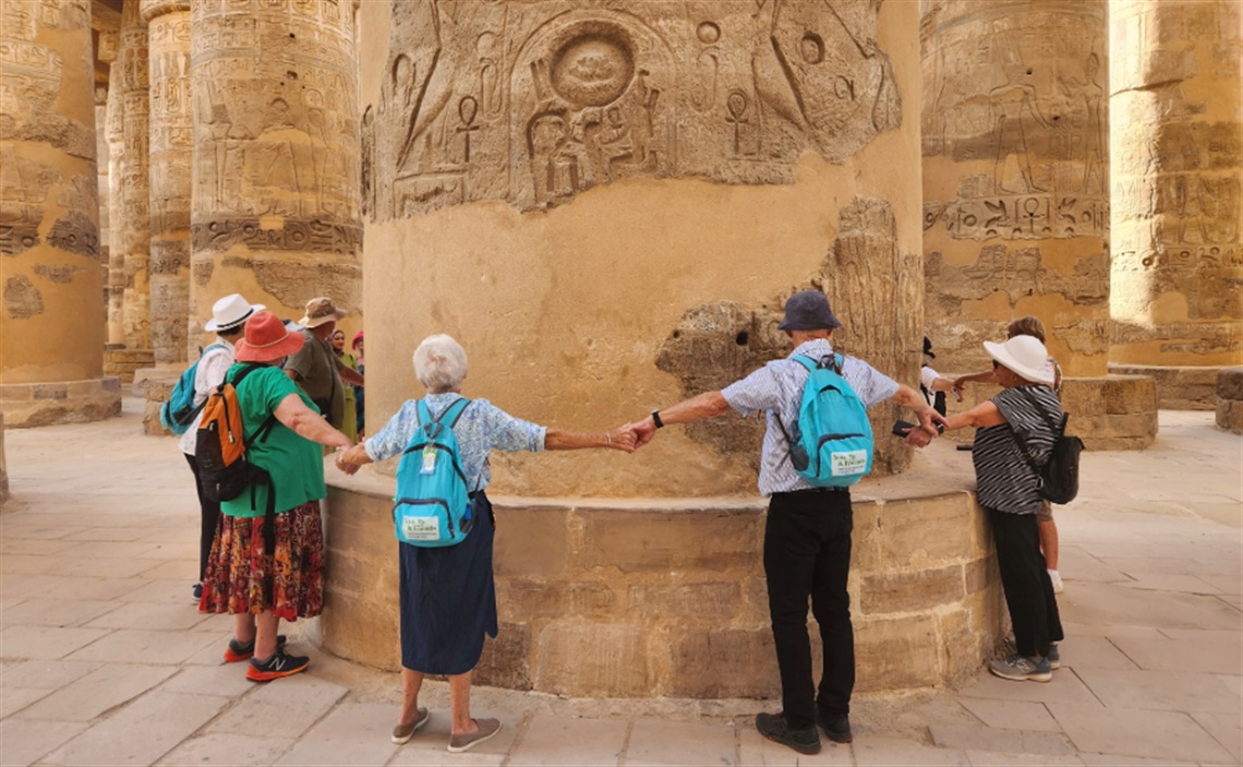 Group of tourists holding hands in a circle around an ancient column with hieroglyphics at an archaeological site. The column features intricate carvings and symbols.