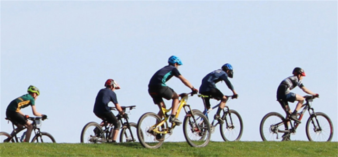 Five individuals riding mountain bikes on a grassy hill under a clear blue sky, all wearing helmets