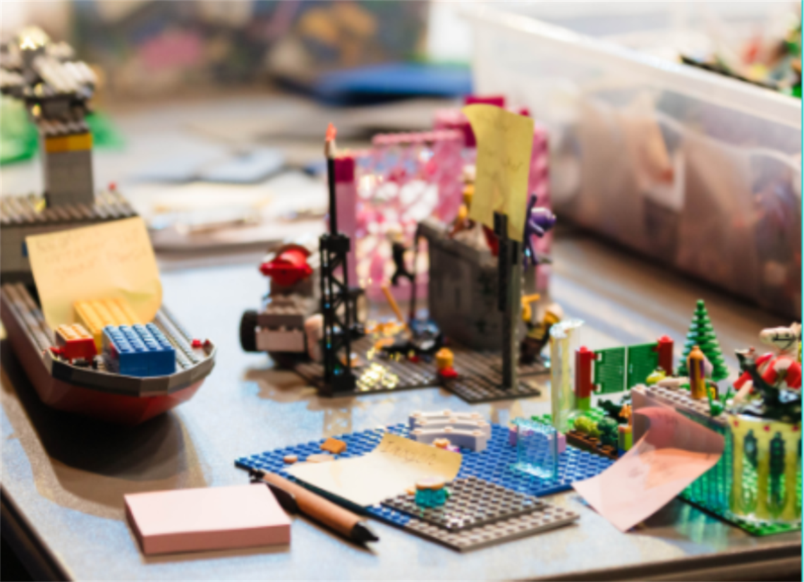 A tabletop covered with colorful LEGO creations, including a boat, small structures, and a green scene with trees. Several pieces have sticky notes attached, and a pencil and pink notepad are placed nearby. A clear plastic bin filled with assorted LEGO pieces is visible in the background.