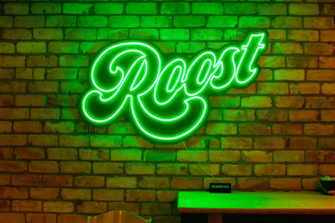 Green neon sign displaying the word “Roost” in a cursive font mounted on a brick wall. Below the sign is a wooden table with a small black “Reserved” sign and a plant in a pot.