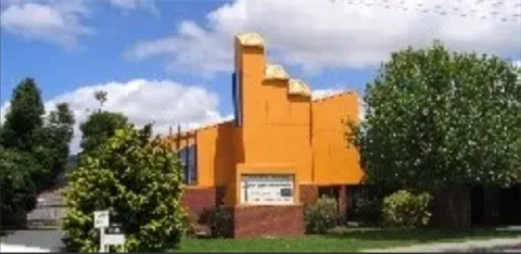 Bright orange building with three protruding roof structures, surrounded by trees and bushes. A sign is positioned in front of the building, though its text is unclear. The sky is partly cloudy with visible blue patches.