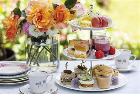 Three tier afternoon tea stand with finger sandwiches, scones, cake slices, macarons and berry desserts beside a vase of colourful roses on a sunlit outdoor table.