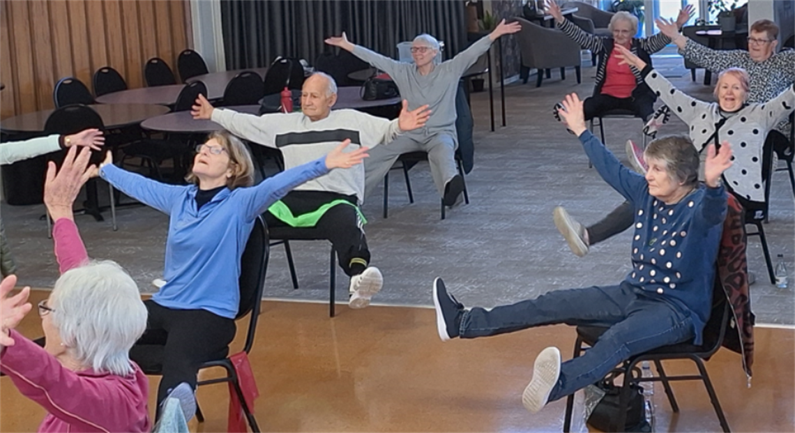Group of people seated in a room with wooden walls, participating in an exercise or stretching activity with arms and legs extended; tables and chairs are visible in the background