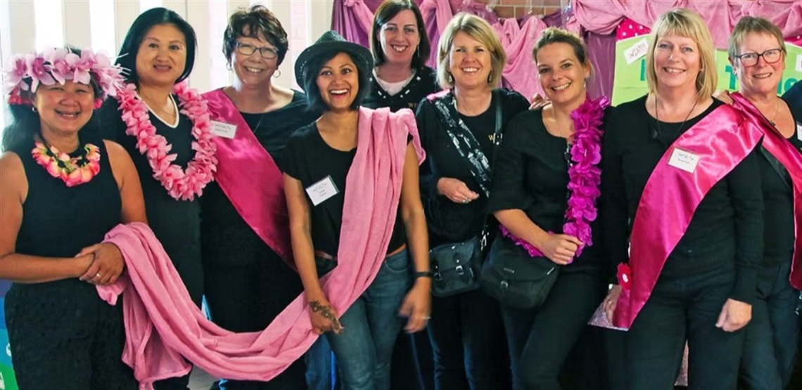 Group of nine people dressed in black with pink accessories including sashes, scarves, and leis, posing in front of a pink fabric backdrop at an event. Some wear name tags, suggesting a formal or themed gathering