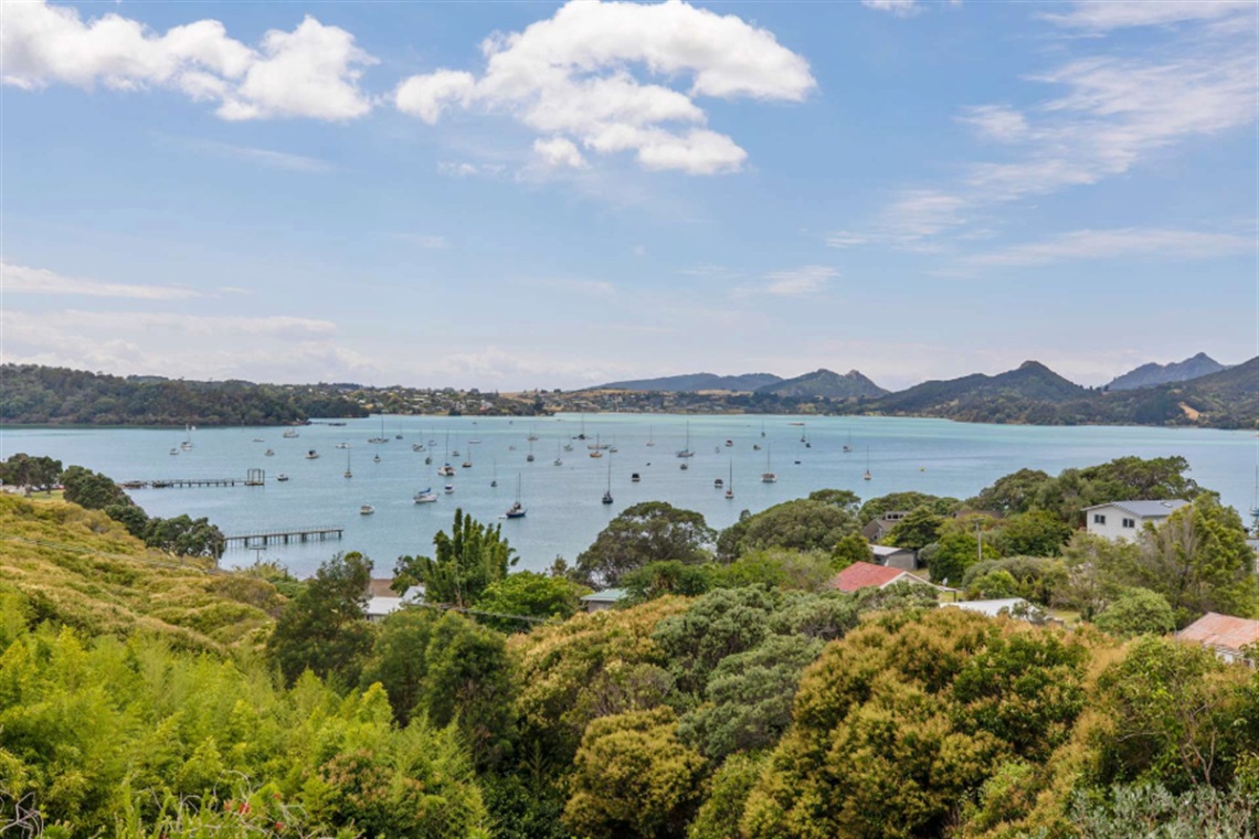 Scenic coastal view of Parua Bay in Northland, New Zealand. The image shows calm blue water dotted with moored sailboats, surrounded by lush green hills and trees. In the foreground are residential rooftops partially hidden by dense vegetation, while the background features rolling hills and a partly cloudy sky.
