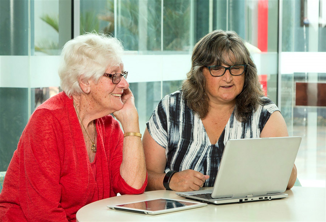 An elderly woman with a tablet on a table sitting with a librarian looking at a laptop screen. 