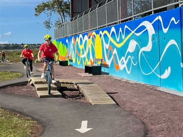 Bikers riding on wooden steps with mural in the background on beginner skills track at Bike Park on Pohe Island. The colourful mural next to the beginners area was created by artist Lucinda Skye.
