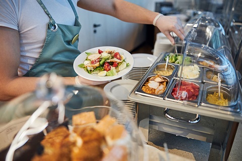 A person in a light blue apron holds a salad plate at a buffet with various dishes in metal containers. 