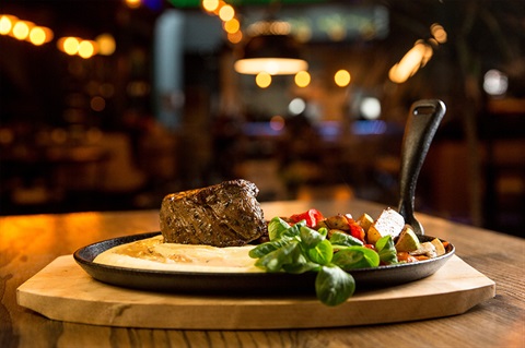 A wooden table with a cast iron skillet containing a cooked steak, mashed potatoes, and a side of vegetables including greens and red bell peppers. The background is blurred with warm lighting, suggesting a cozy restaurant setting. 