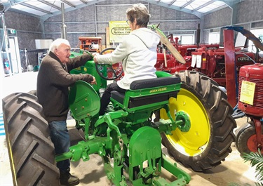 An older adult lady sitting on a tractor with an older adult man standing nearby.