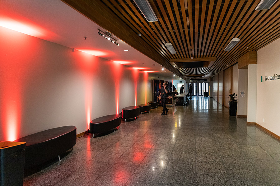The Forum North concourse features a polished stone floor and wooden slatted ceiling. Red lights illuminate the left wall with black cushioned benches, while people gather near glass doors at the far end. 