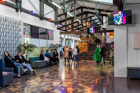 People gather in the Forum North foyer, socialising before an event beneath exposed beams and display screens. 